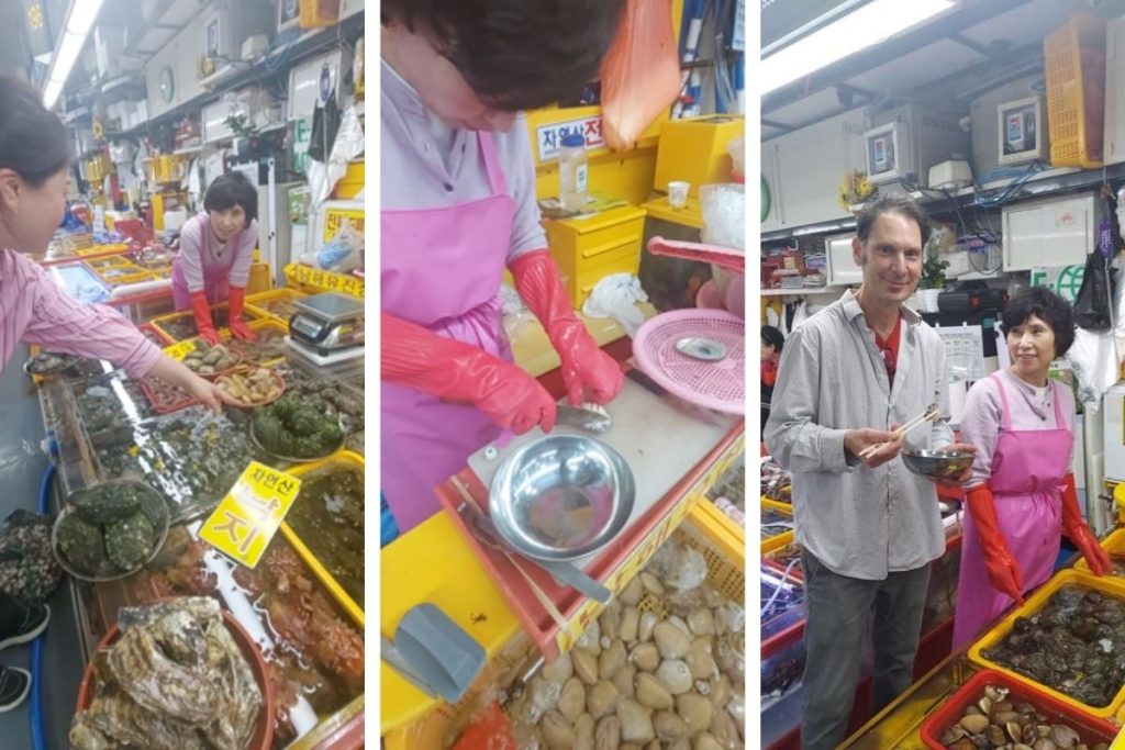 A seafood vendor at Jagalchi Market in Busan prepares fresh abalone. A traveler enjoys a bite while chatting with the vendor. Fresh seafood everywhere.