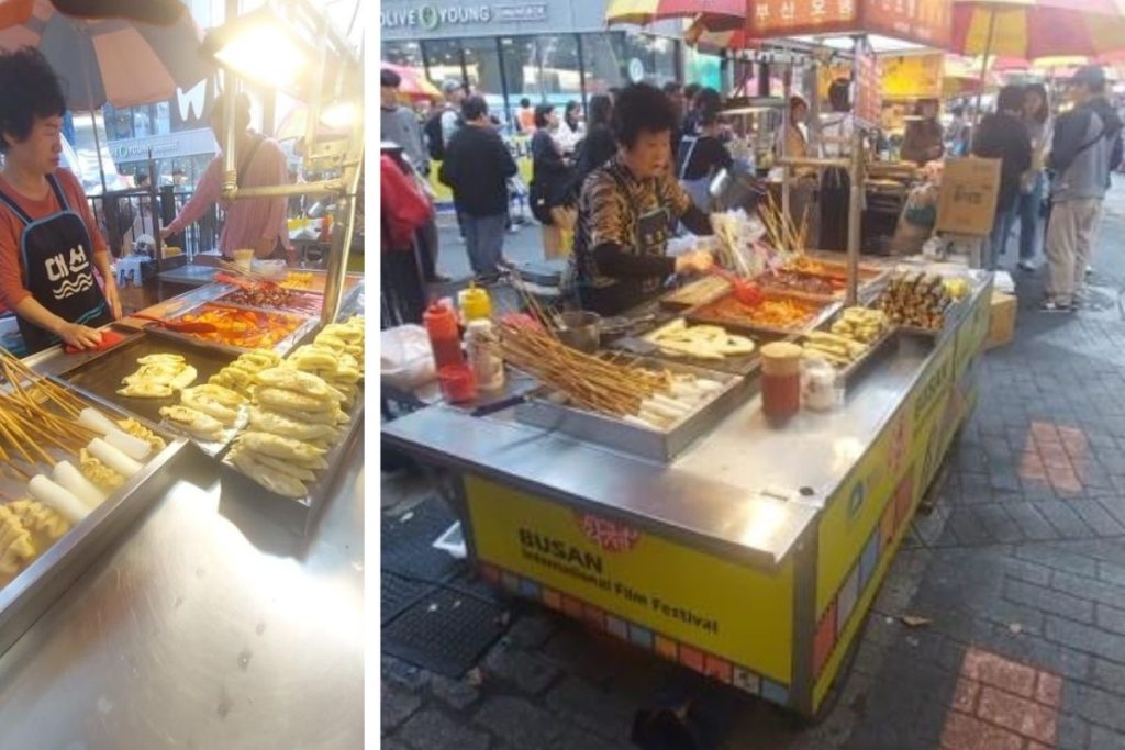 Vendors at Gukje Market, grill dumplings, skewers, and rice cakes. A lively street food scene with sizzling flavors and colorful umbrellas.